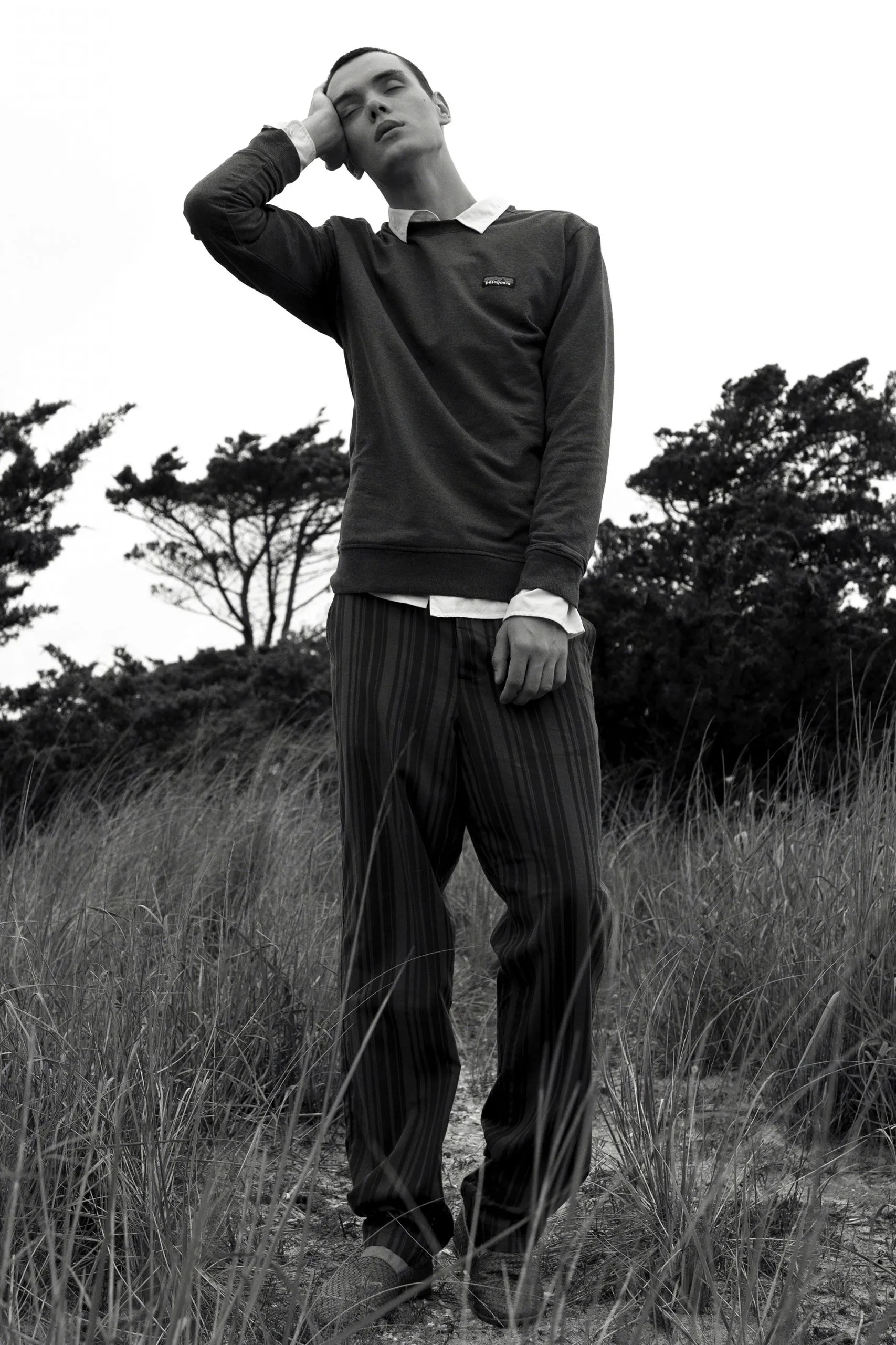Black and white photograph of a man in dark trousers and a long-sleeve top standing in tall grass, hand to his head, wind in the field