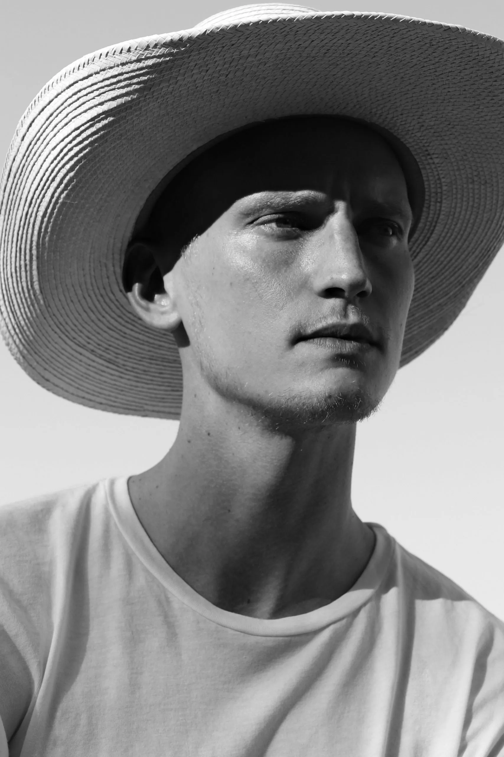 Black and white photograph of a man in a wide straw hat looking up, photographed from below against a pale sky