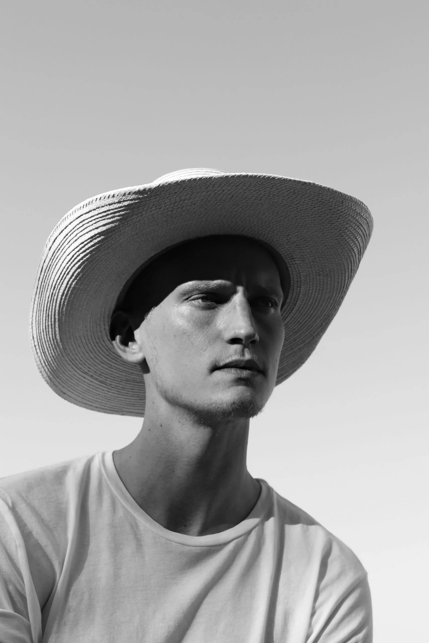 Black and white photograph of a man in a wide straw hat and white tee, standing in an open field