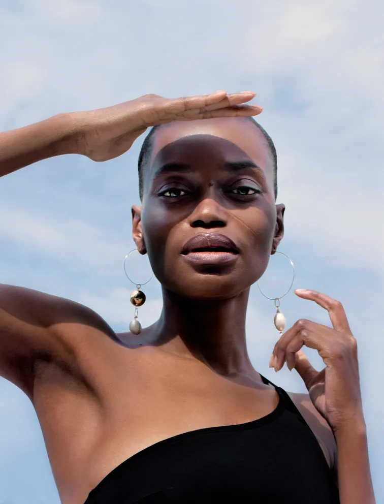 Model in a black one-shoulder top shielding her eyes from the sun against a blue sky